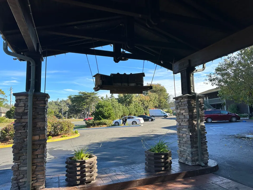 A stone-pillared covered entrance with a hanging sign, planters and a car park under a bright blue sky.