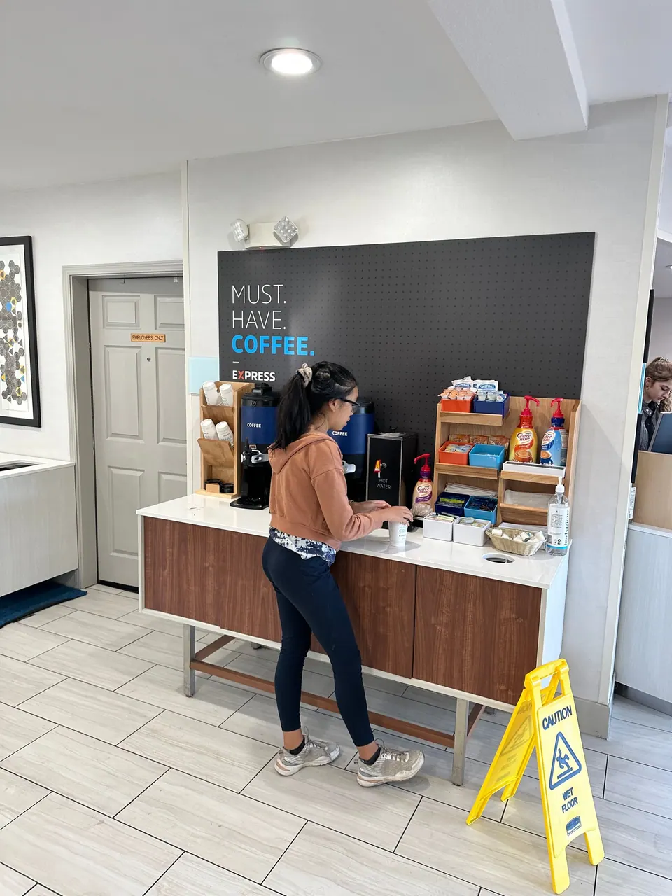 A girl in a brown hoodie pours coffee at a lobby counter under a wall sign reading Must. Have. Coffee.