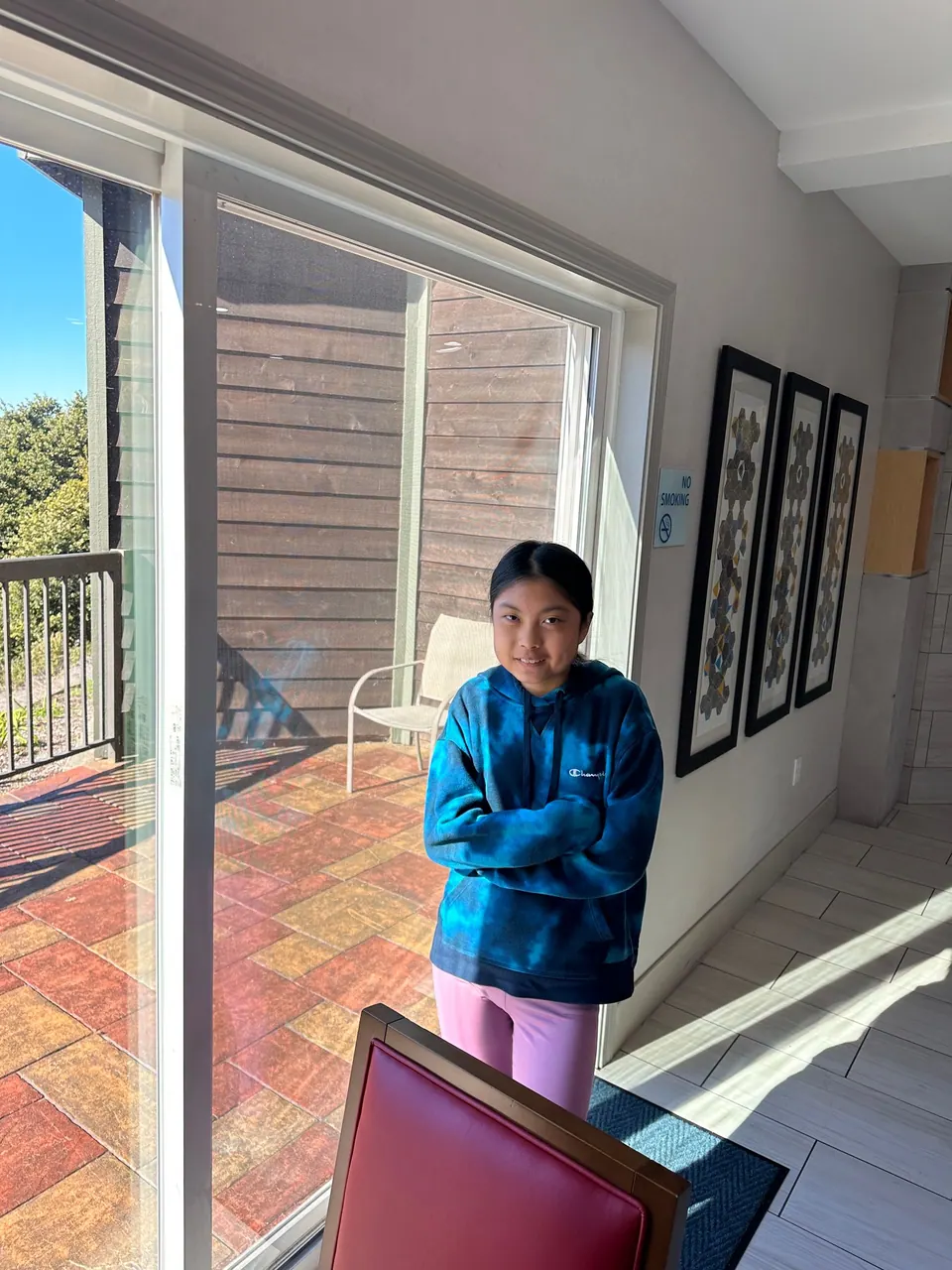 A girl in a blue tie-dye hoodie stands with her arms crossed beside a glass door to a balcony, a red chair in the corner.