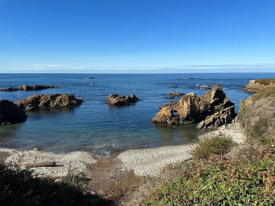 Blue water, dark rocks and a gravel cove below a grassy bluff under a cloudless sky.