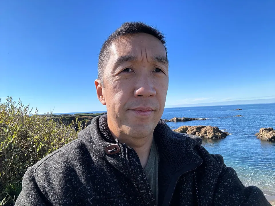 A man in a gray fleece takes a selfie on a coastal bluff with the sea and rocks behind him.