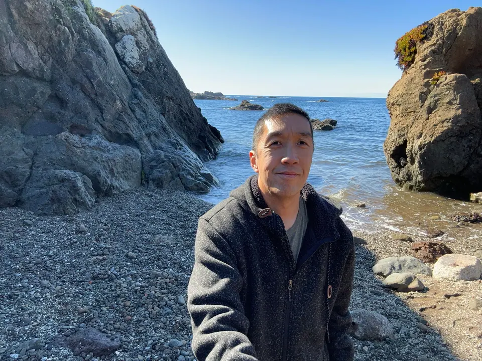 A man in a gray fleece stands on a beach of gray pebbles between two rock walls with the sea behind him.