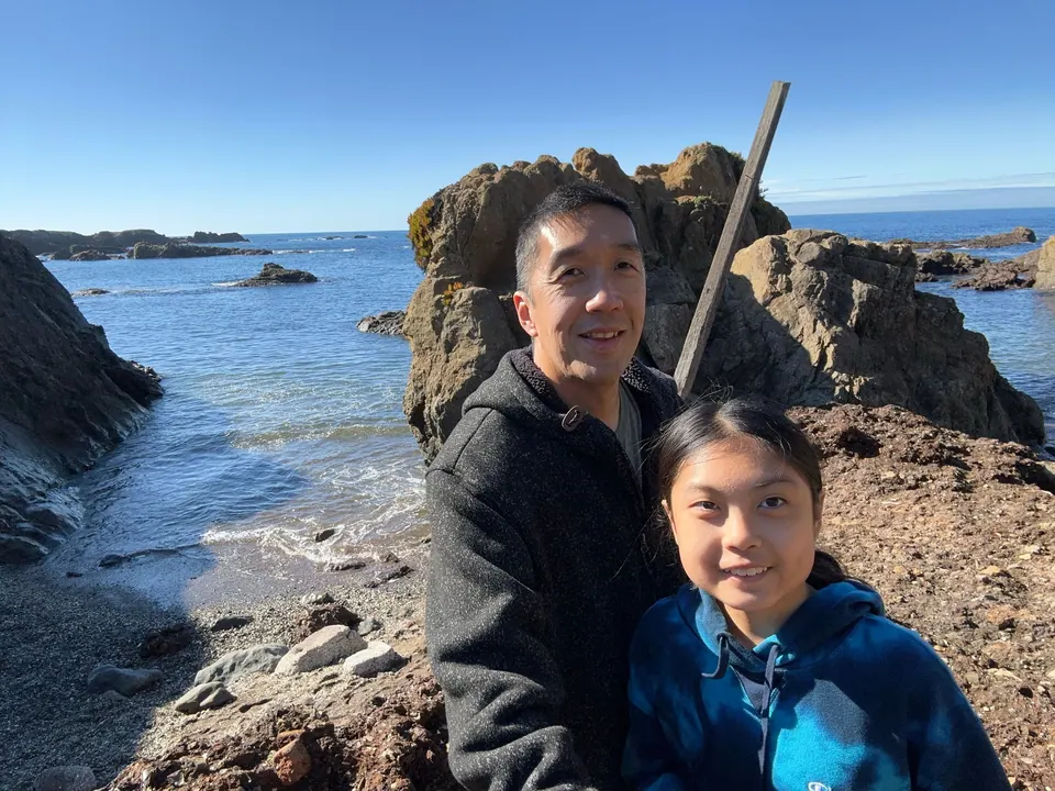 A man and a girl in a blue hoodie lean together in a selfie, a rock wall and calm water behind them.