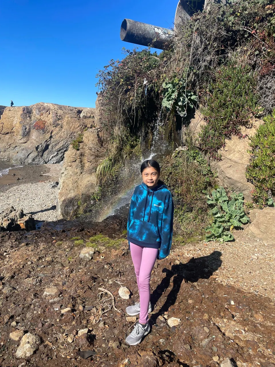 A girl in a blue tie-dye hoodie and pink leggings stands on brown dirt below a green bank with a small trickle of water and a rusty pipe.