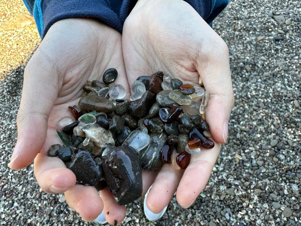 Two cupped hands hold smooth sea glass and dark wet pebbles over a gray gravel beach.