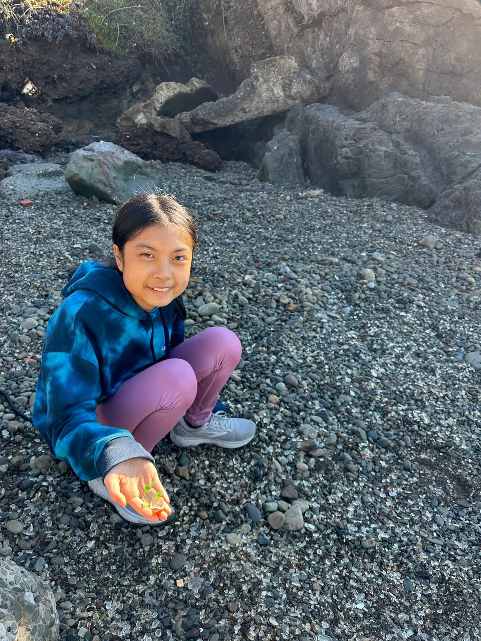 A girl in a blue tie-dye hoodie and lilac leggings crouches on gray gravel, smiling, holding glass in her palm.