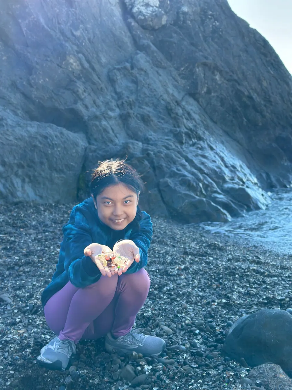 A crouching girl holds out both hands with small pieces of glass in front of a dark cliff and the sea.