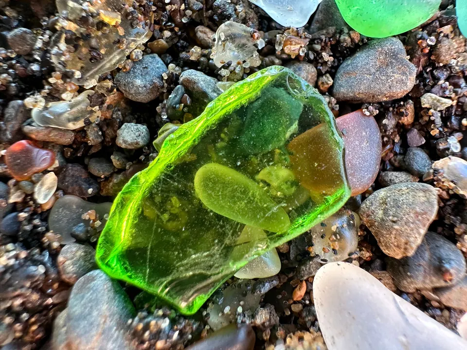 A large translucent green chunk of glass sits among pebbles and frosted glass in close-up.