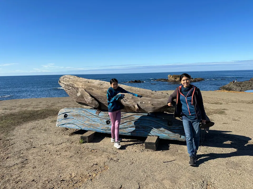 A girl and a woman lean against a big driftwood whale carving and bench above the sea.