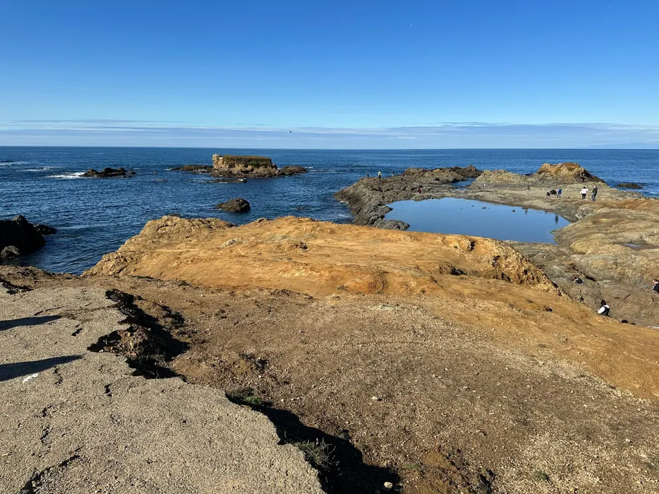 Rounded golden rock ledges, a still tide pool and rock stacks under a clear blue sky with the Pacific beyond.