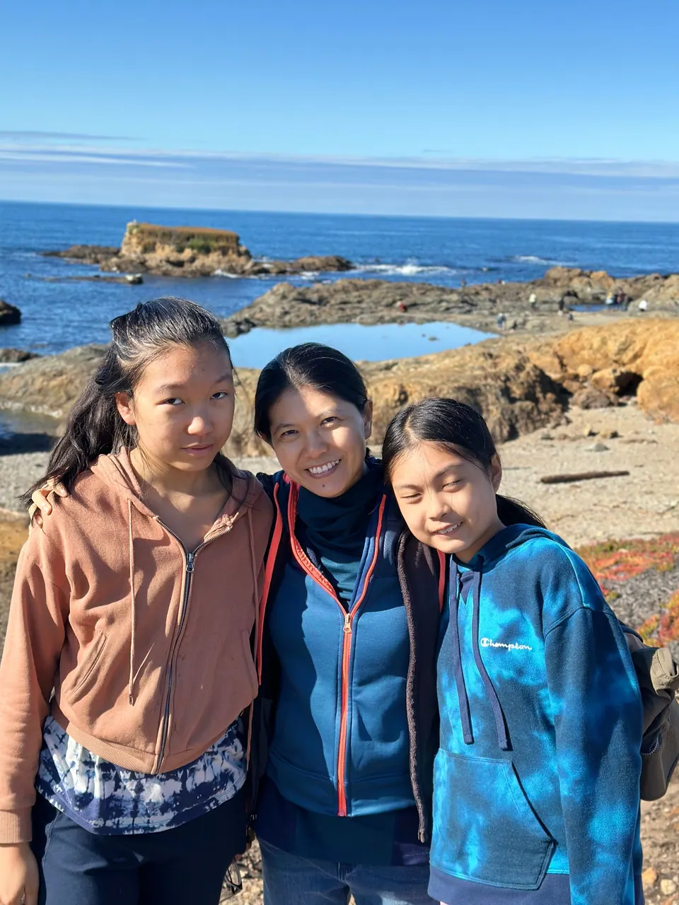 A girl in a brown hoodie, a woman and a girl in a blue tie-dye hoodie stand with their arms around each other above a tide pool and the sea.