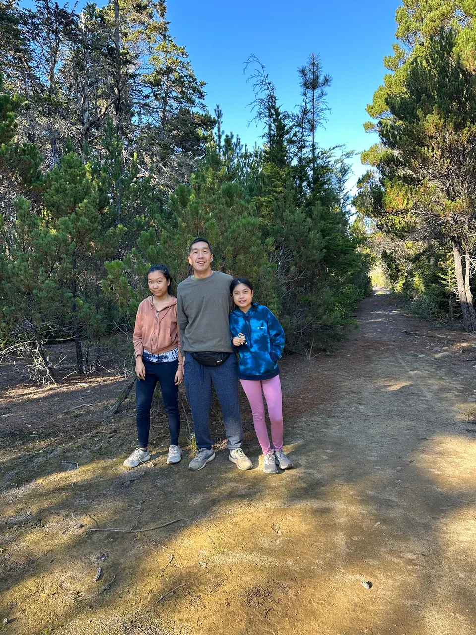 Two girls and a man stand on a pale sandy trail lined with stunted pines and cypress.