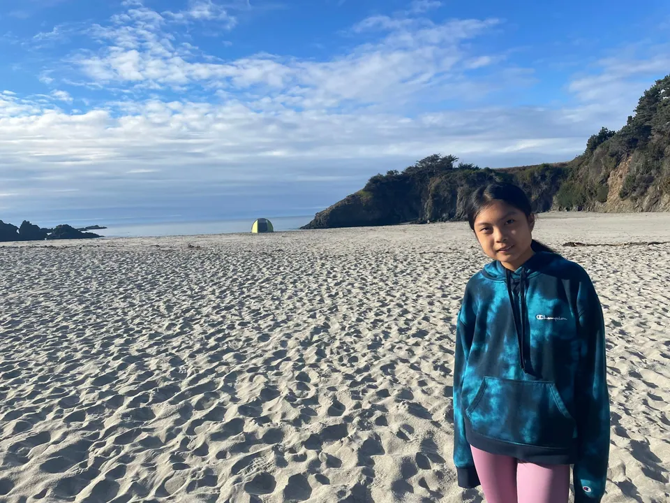 A girl in a blue tie-dye hoodie stands on a wide, trampled beach with a green headland and the sea behind.