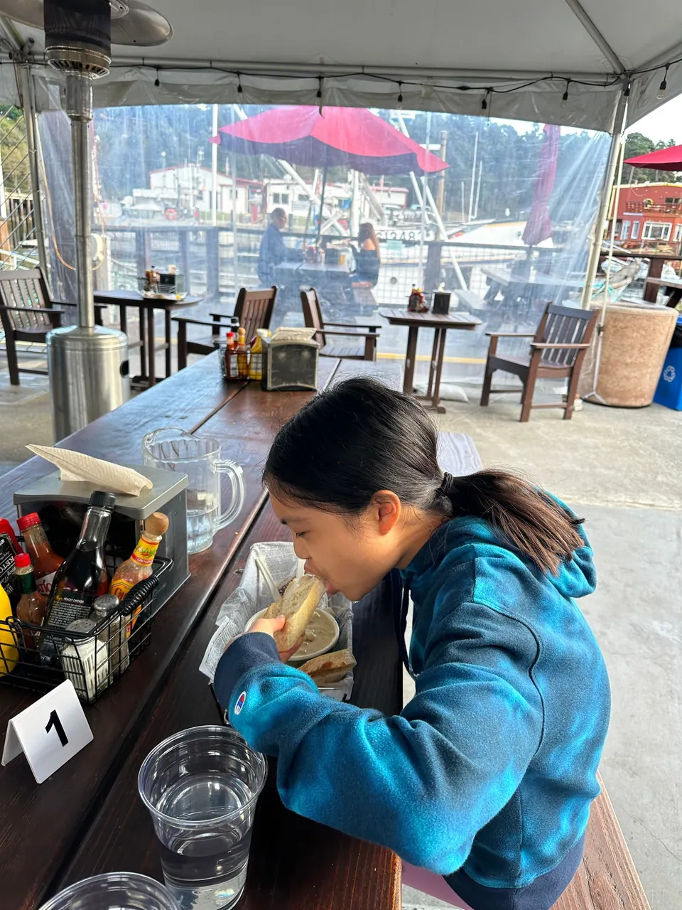 A girl in a blue tie-dye hoodie eats at a counter under a white tent with a glass of water and condiments, the harbor behind.