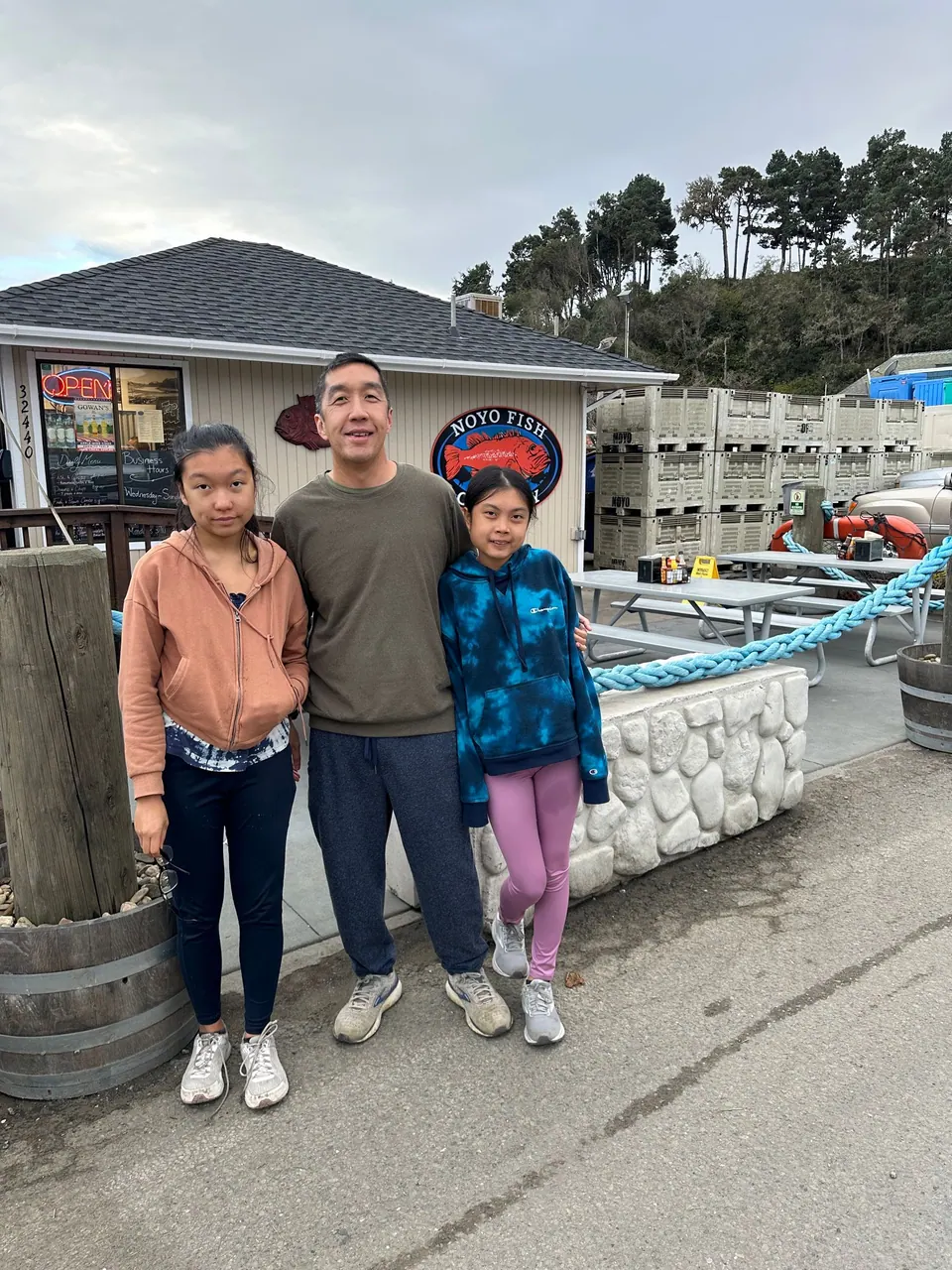 A girl, a man and a girl stand in front of a fish market with a red rockfish sign and a stack of fish crates behind.