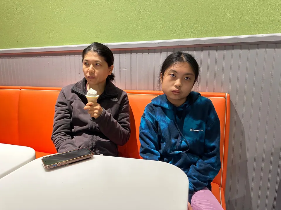 A woman holds a cone and a girl in a blue hoodie sits beside her on an orange bench in a bright ice cream shop.
