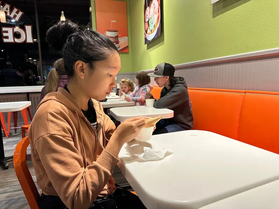 A girl in a brown hoodie eats at a white table under a lime-green wall.
