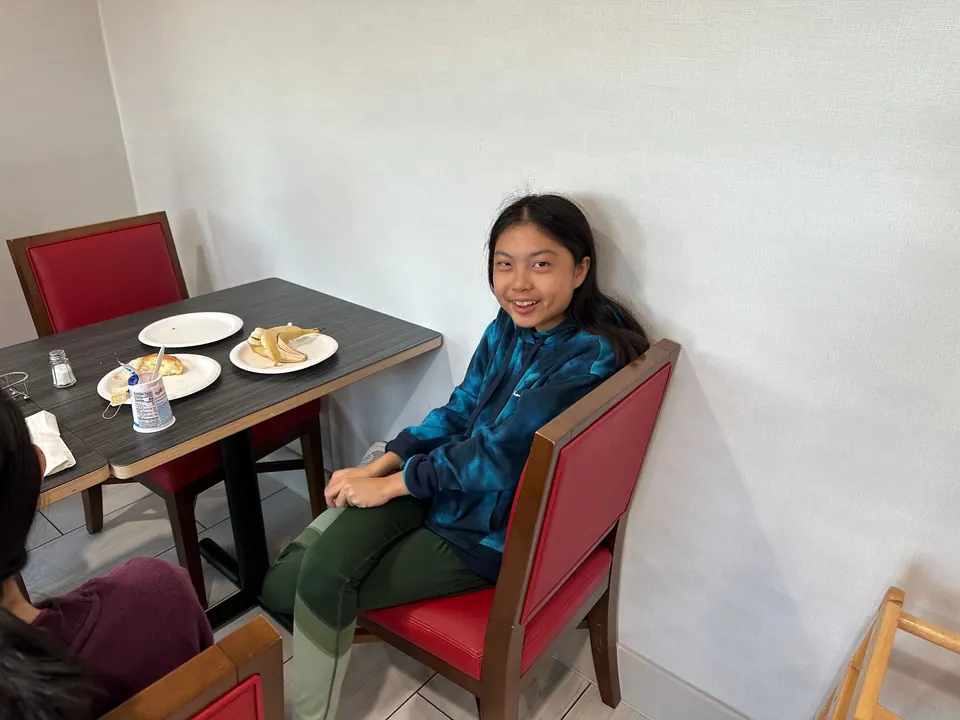 A girl in a blue tie-dye hoodie and green leggings sits smiling on a red chair beside a table with plates of waffle and eggs.