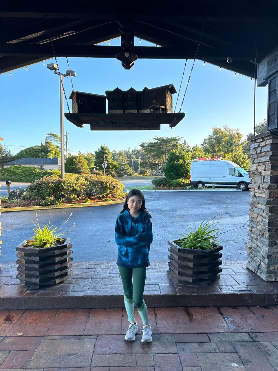 A girl in a blue tie-dye hoodie and teal leggings stands with arms crossed under a covered entrance in bright sun.