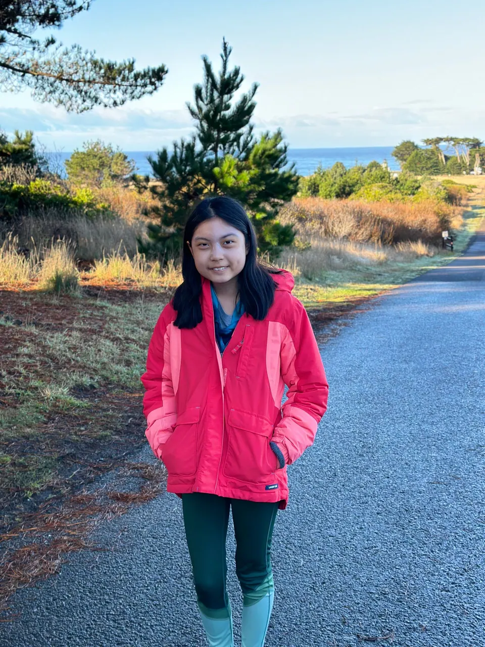 A girl in a bright pink-red jacket and teal leggings stands on a paved road in early sun, a young pine and the sea behind.