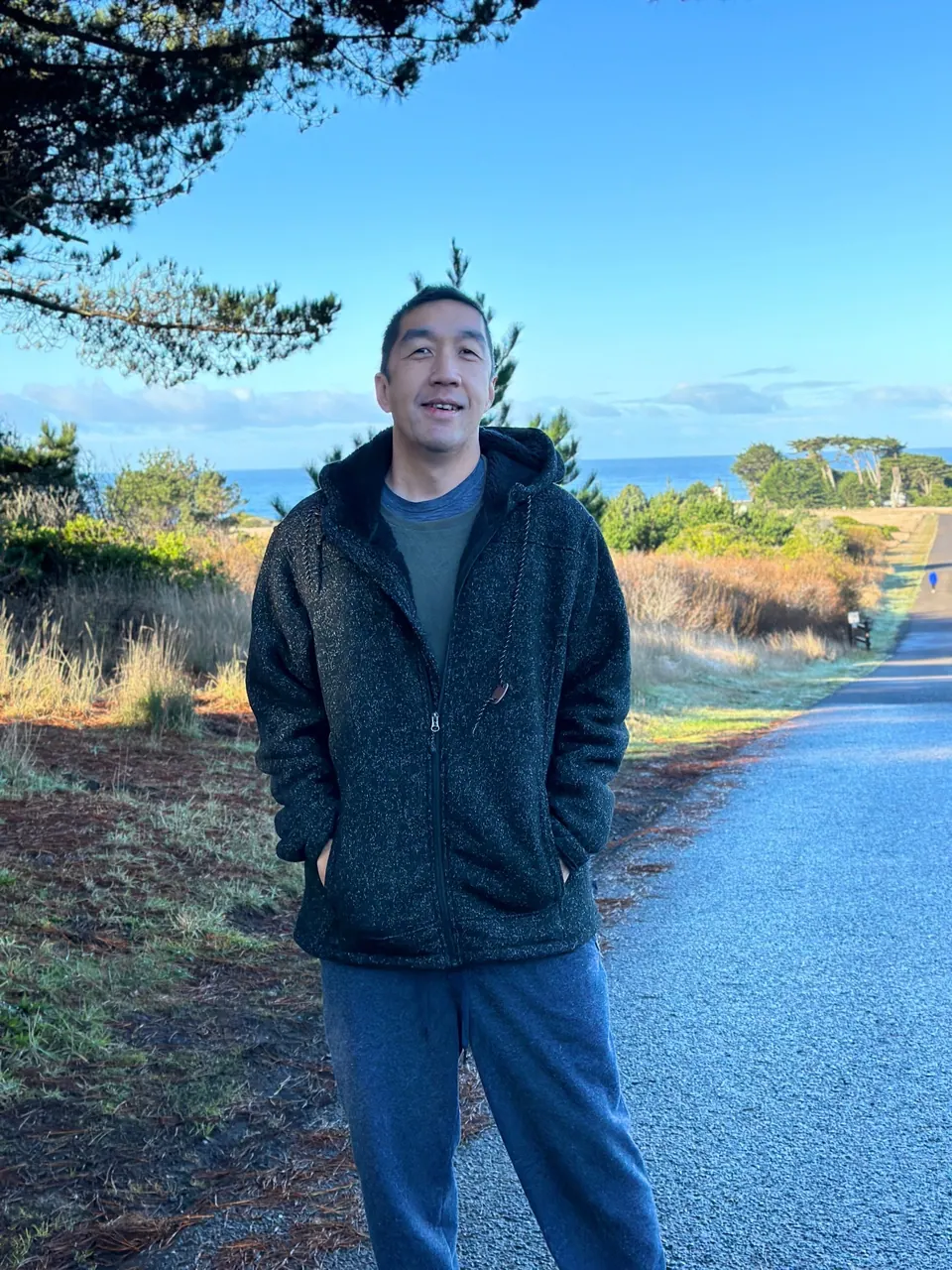 A man in a dark fleece stands on a road in dry grass with pines and the sea behind him.