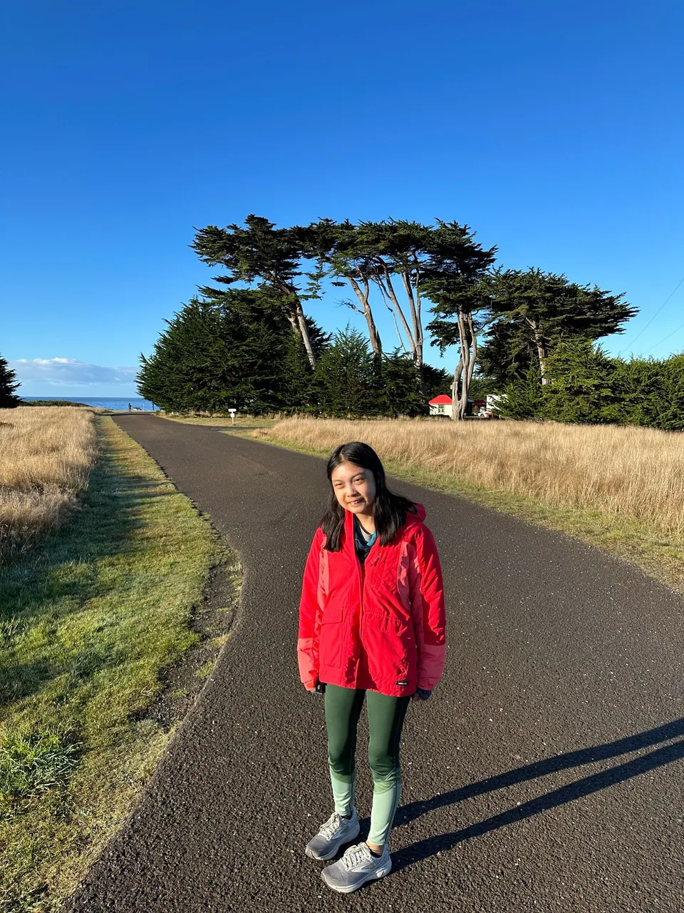 A girl in a red jacket smiles on a grassy road below a windbreak of cypress; a red-roofed house is in the distance.