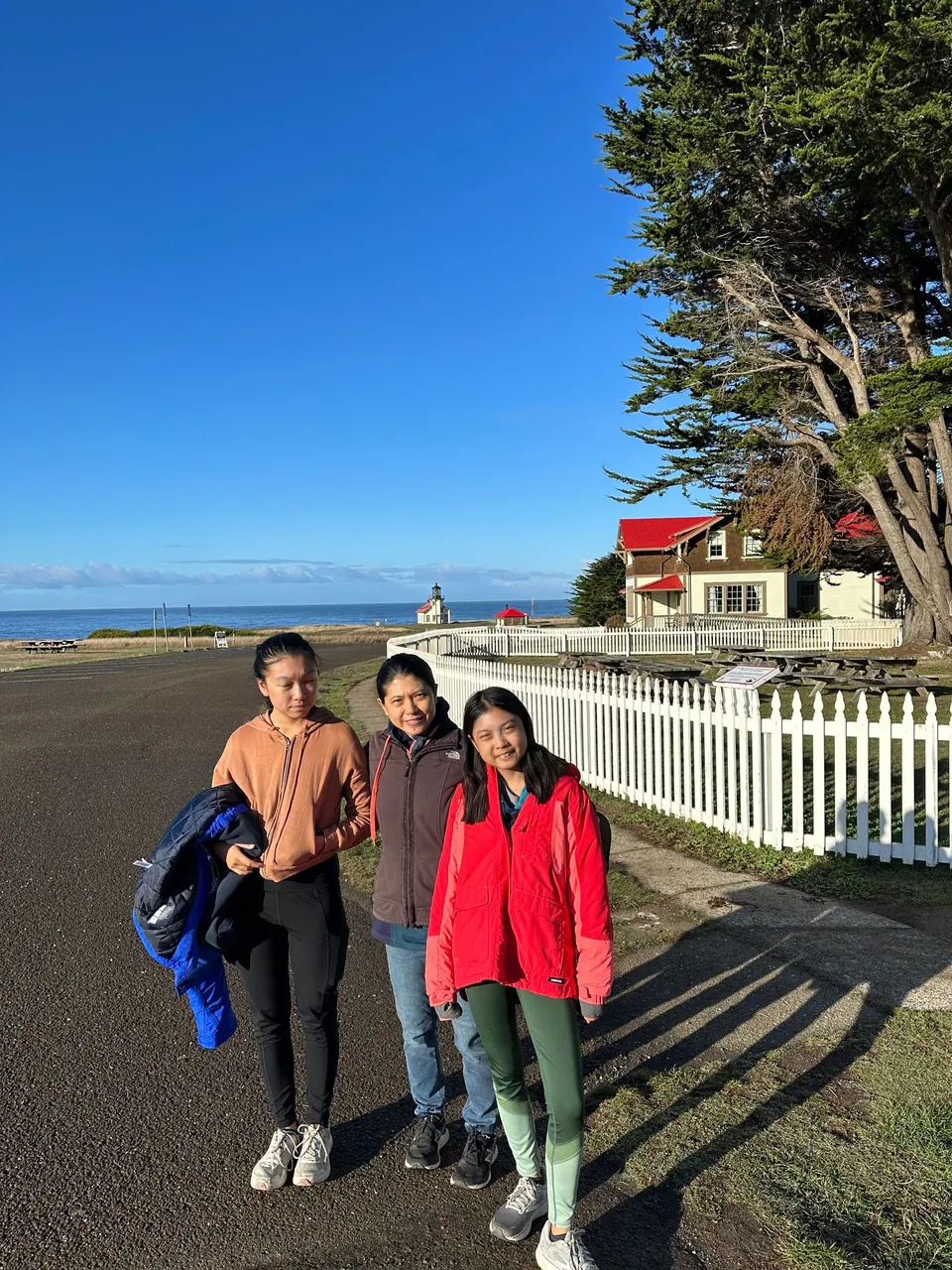 A girl in a tan hoodie carrying a blue coat, a woman in a brown jacket and a girl in a red jacket stand together on a paved road beside a white picket fence, with a red-roofed house and a distant lighthouse behind.