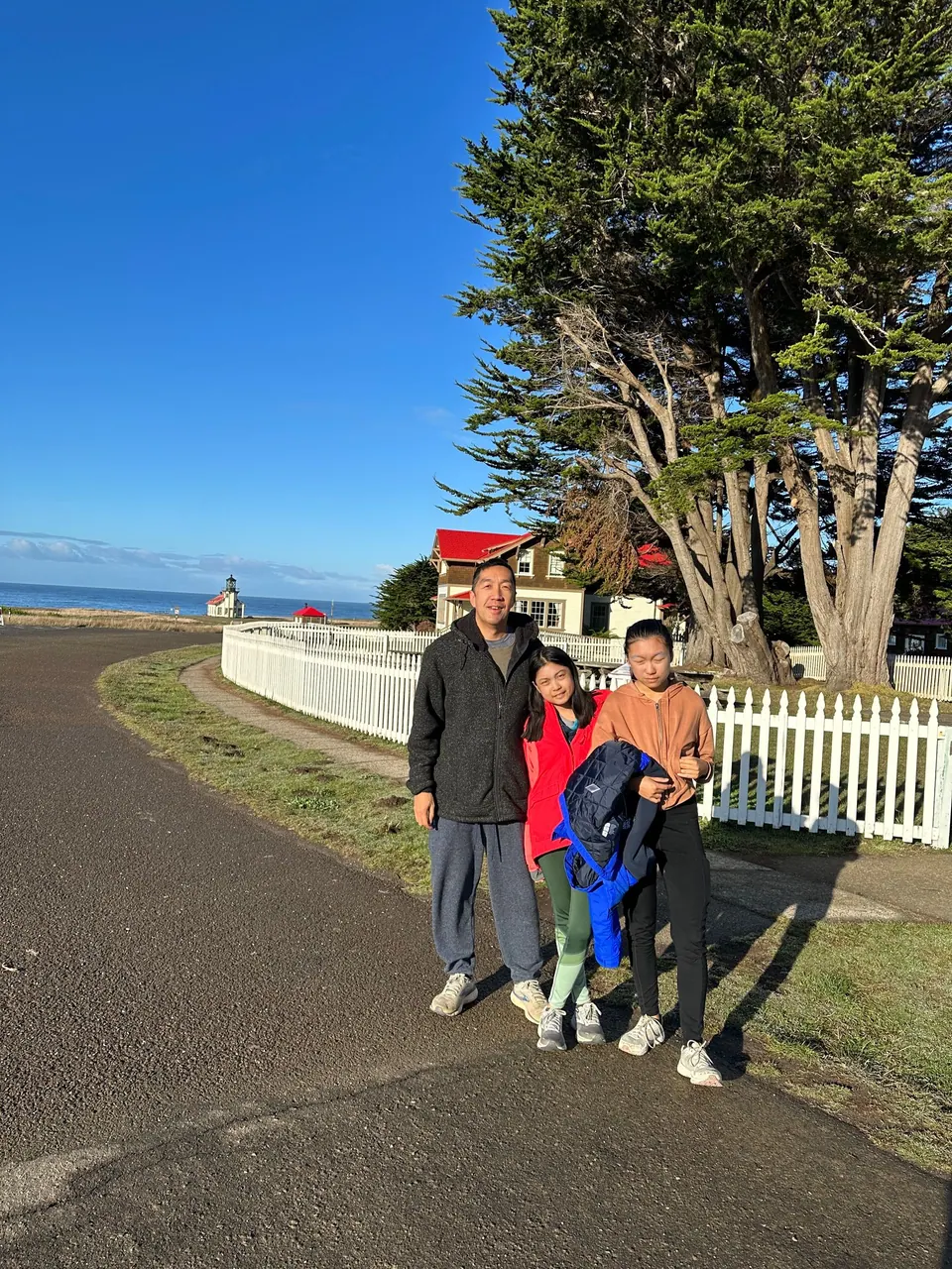 A man, a girl in a red jacket and a girl in a brown hoodie stand in front of the picket fence and a red-roofed house.