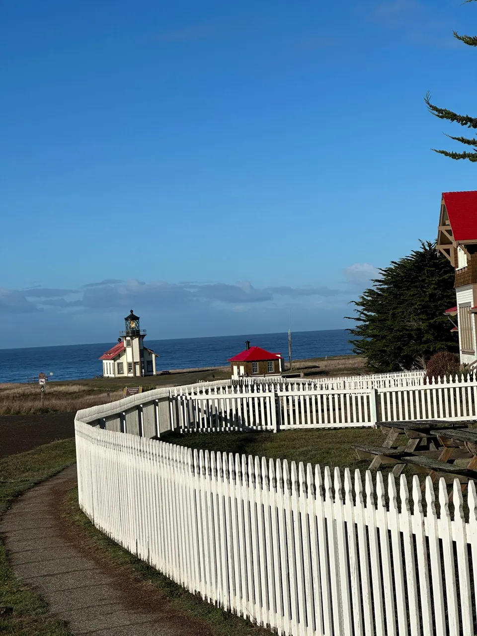 A white picket fence curves along a road toward a red-roofed house and a small lighthouse near the sea.