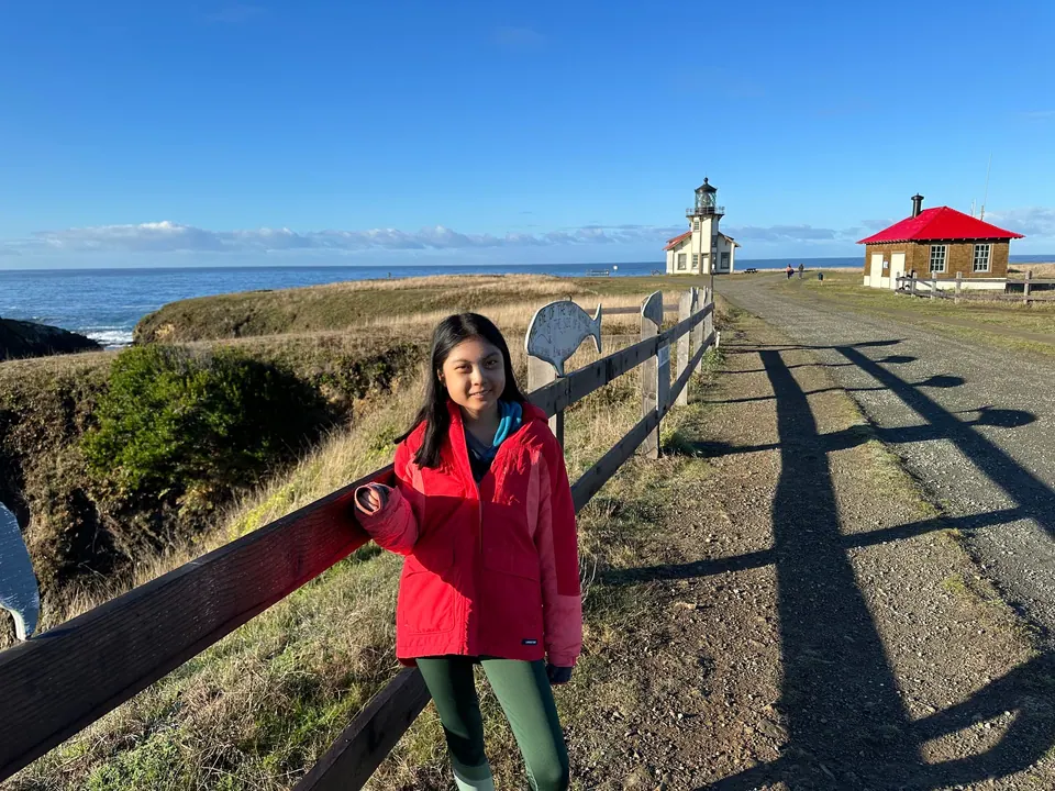 A girl in a red jacket rests on a wooden fence beside a gravel road that leads to a small lighthouse.