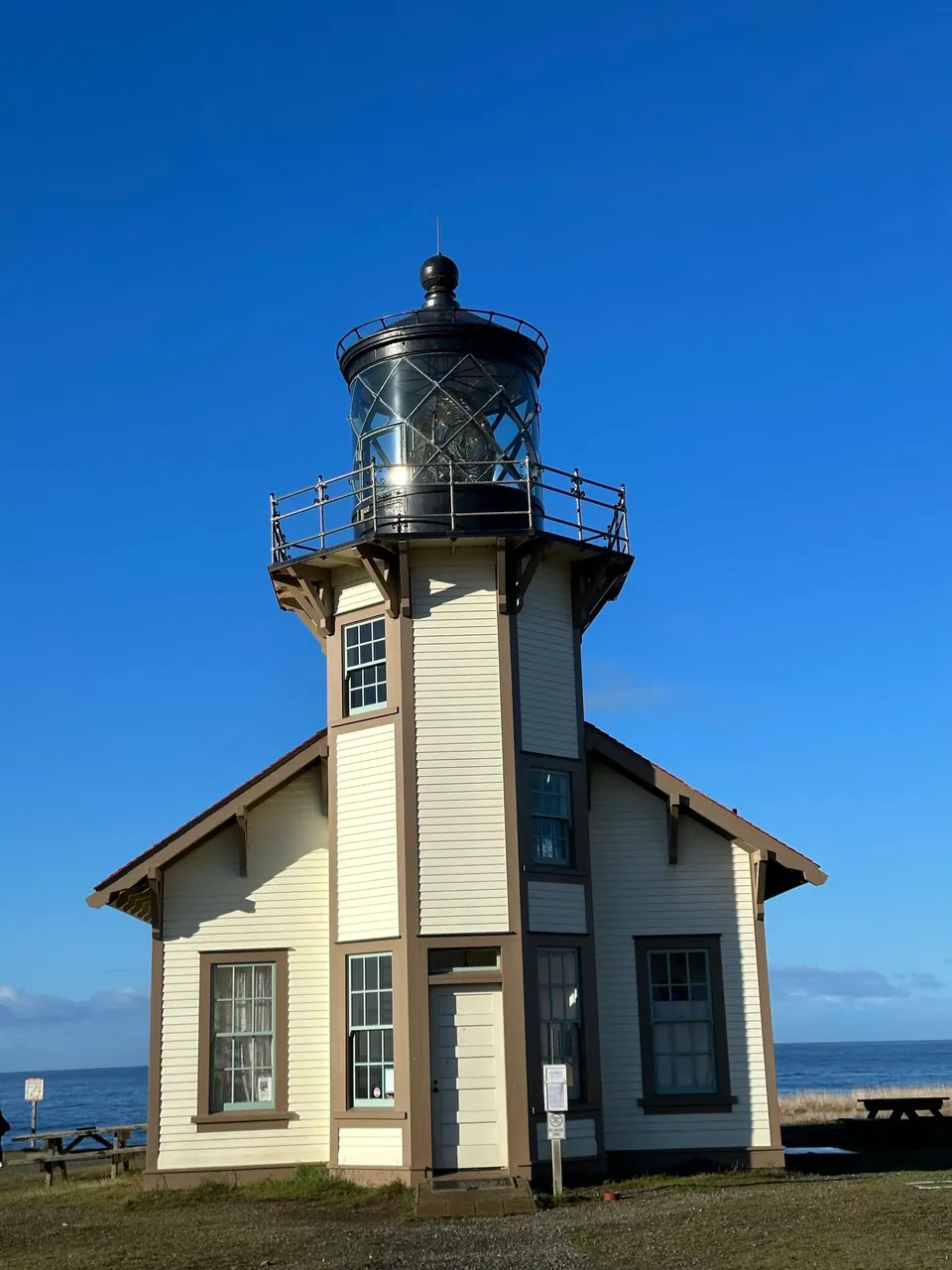 A small white lighthouse tower with a dark lantern room rises from a two-story building, shot from low on the grass against a blue sky.