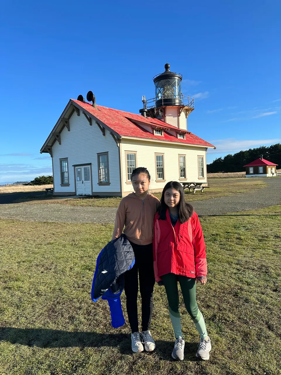 A girl in a light brown hoodie and a girl in a red jacket stand in front of a white building with a red roof and lantern room.
