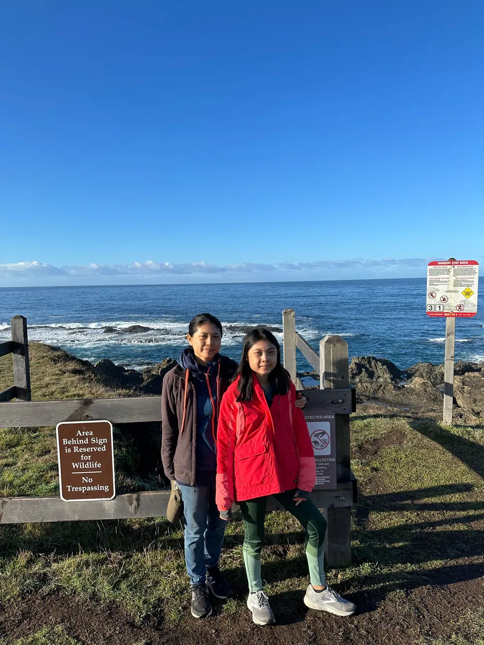 A woman in a dark jacket and a girl in a red jacket stand at a wooden fence beside a brown sign about a wildlife area, with the blue sea behind.