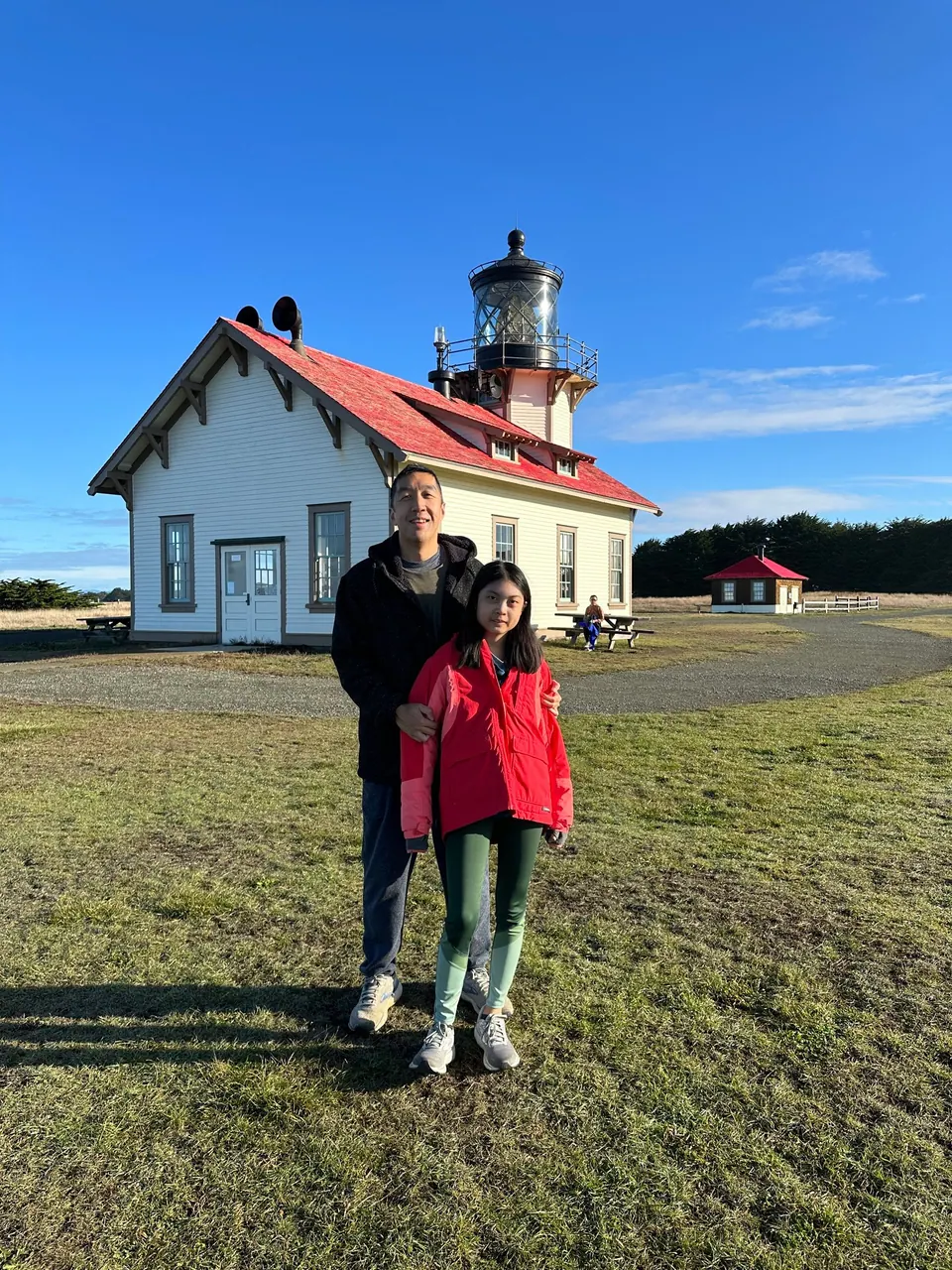 A man and a girl in a red jacket stand in front of the red-roofed lighthouse building.