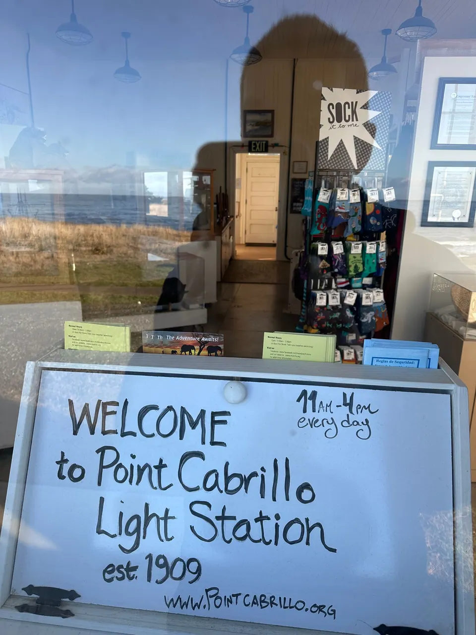 A whiteboard in a window reads Welcome to Point Cabrillo Light Station, est. 1909, open 11 to 4 every day, with shelves of souvenirs behind.