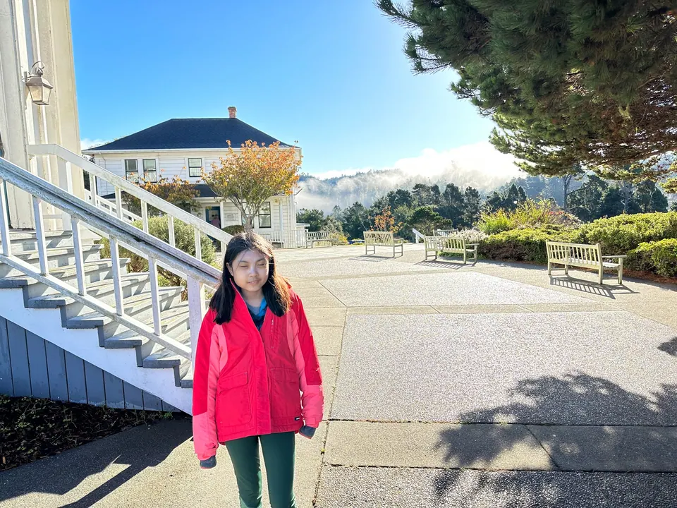 A girl in a red jacket stands at the bottom of white steps with a plaza and a house behind her.