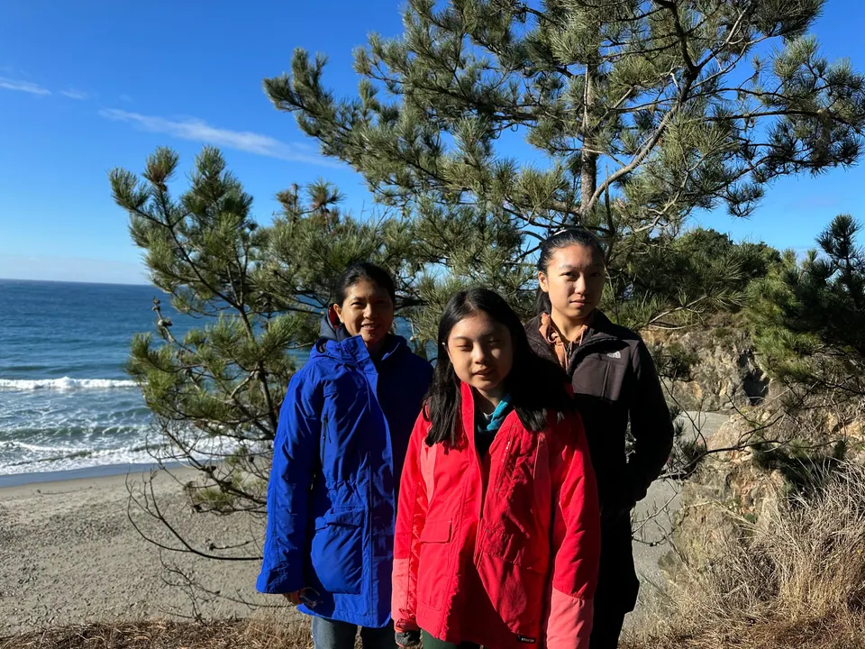 A girl in a red jacket, a woman in blue and a girl in a gray fleece stand at a bluff edge with a wide beach and surf behind.