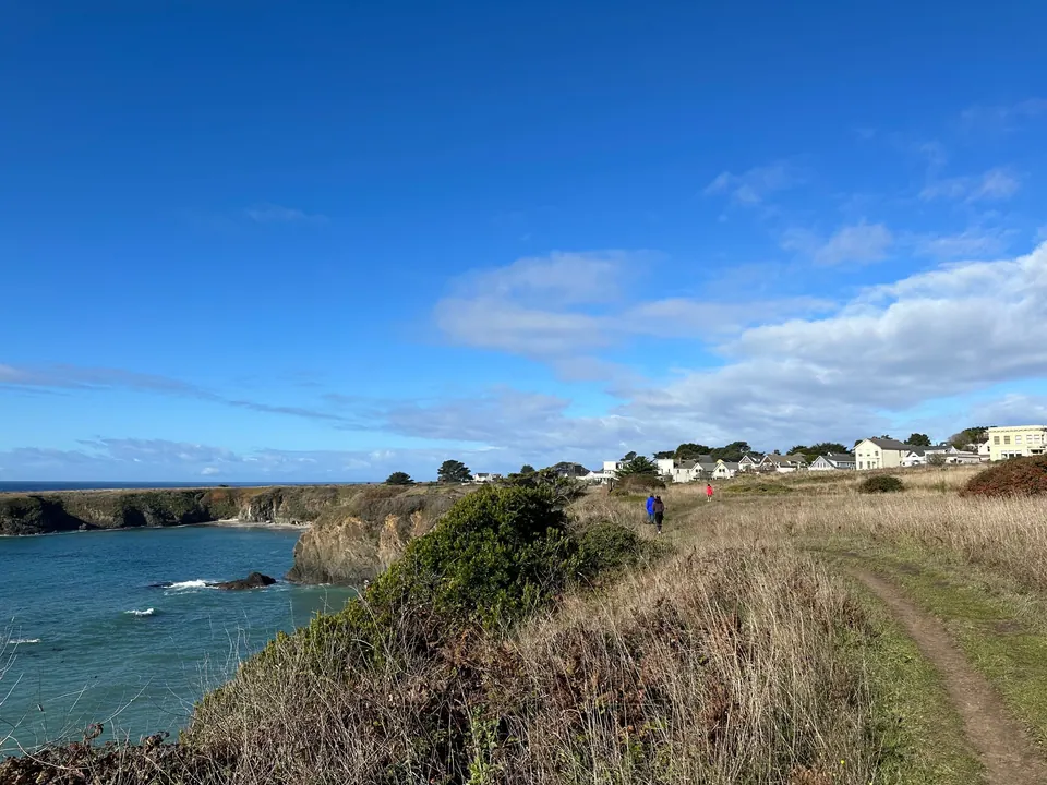 A dirt trail runs above a blue cove toward white houses on a distant bluff.