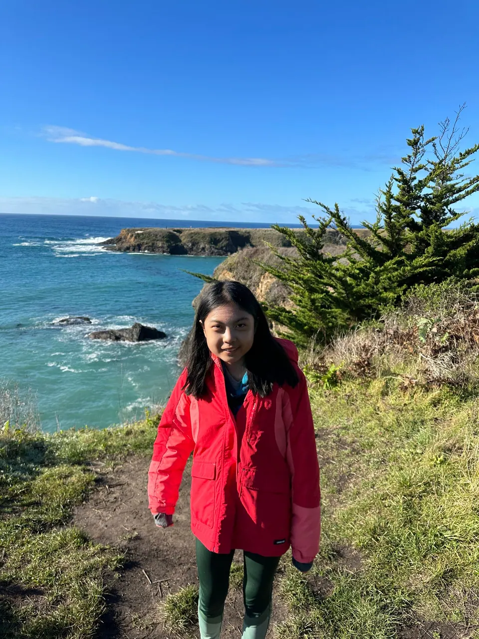 A girl in a red jacket smiles on a bluff with a turquoise cove and a cypress behind.