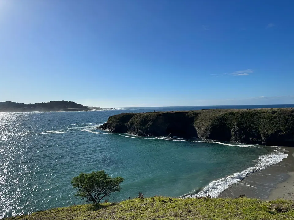 A cove of blue-green water curls onto a small beach below a rocky headland and a horizon of sea.
