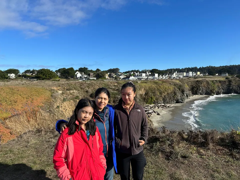 A girl in red, a woman in blue and a girl in a gray fleece stand on a headland with the cove and the village behind.