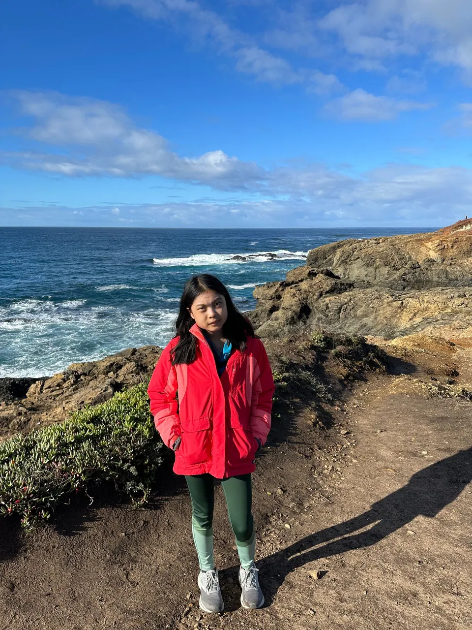 A girl in a red jacket smiles on a rocky bluff over breaking waves.
