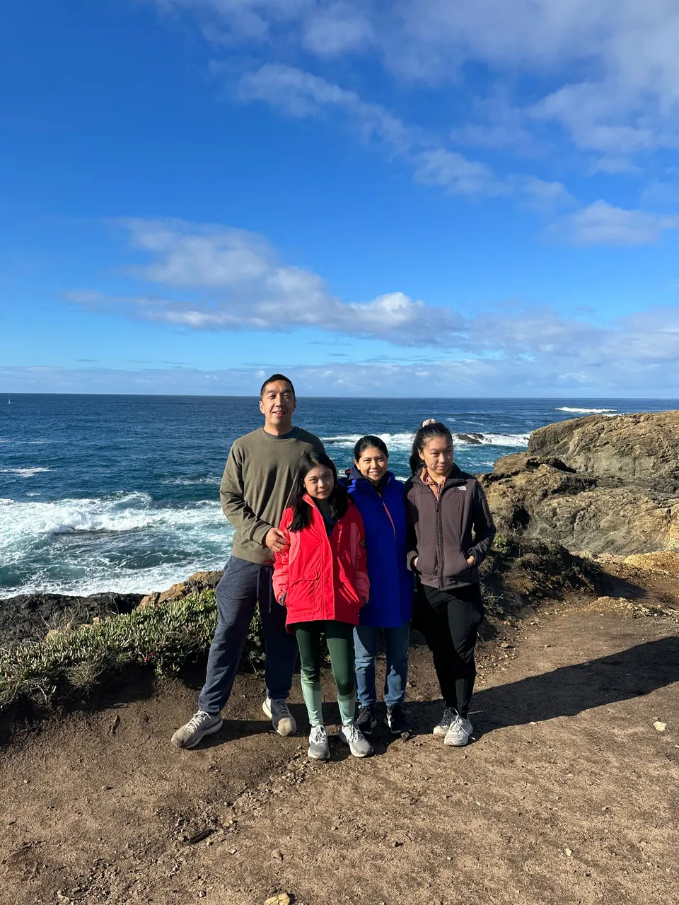 A man, a girl in red, a woman in blue and a girl in a gray fleece stand together on a golden rock bluff with white surf and clouds behind.