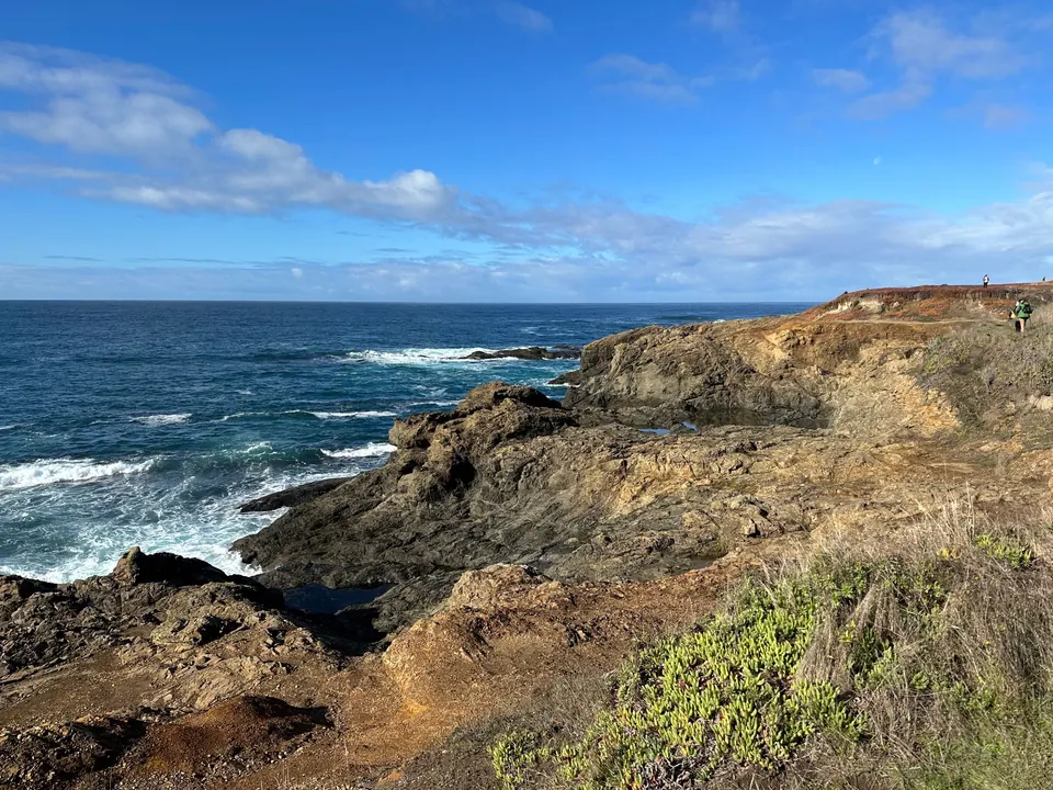 Waves crash against golden, layered rock under a blue sky with drifting clouds.