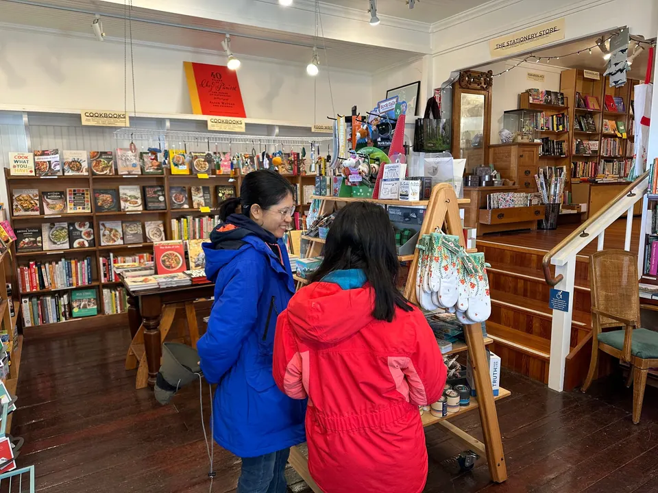 A woman in a blue jacket and a girl in red stand at a table of books, with stairs and bookshelves behind them.