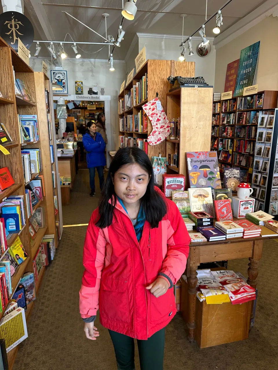 A girl in a red jacket stands among book displays, red stockings hanging from tall shelves.