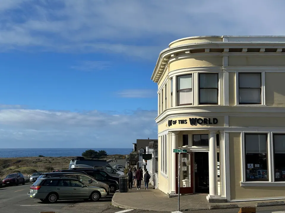 A cream-colored building with a curved corner and a sign reading Out of This World, with parked cars and the ocean beyond.