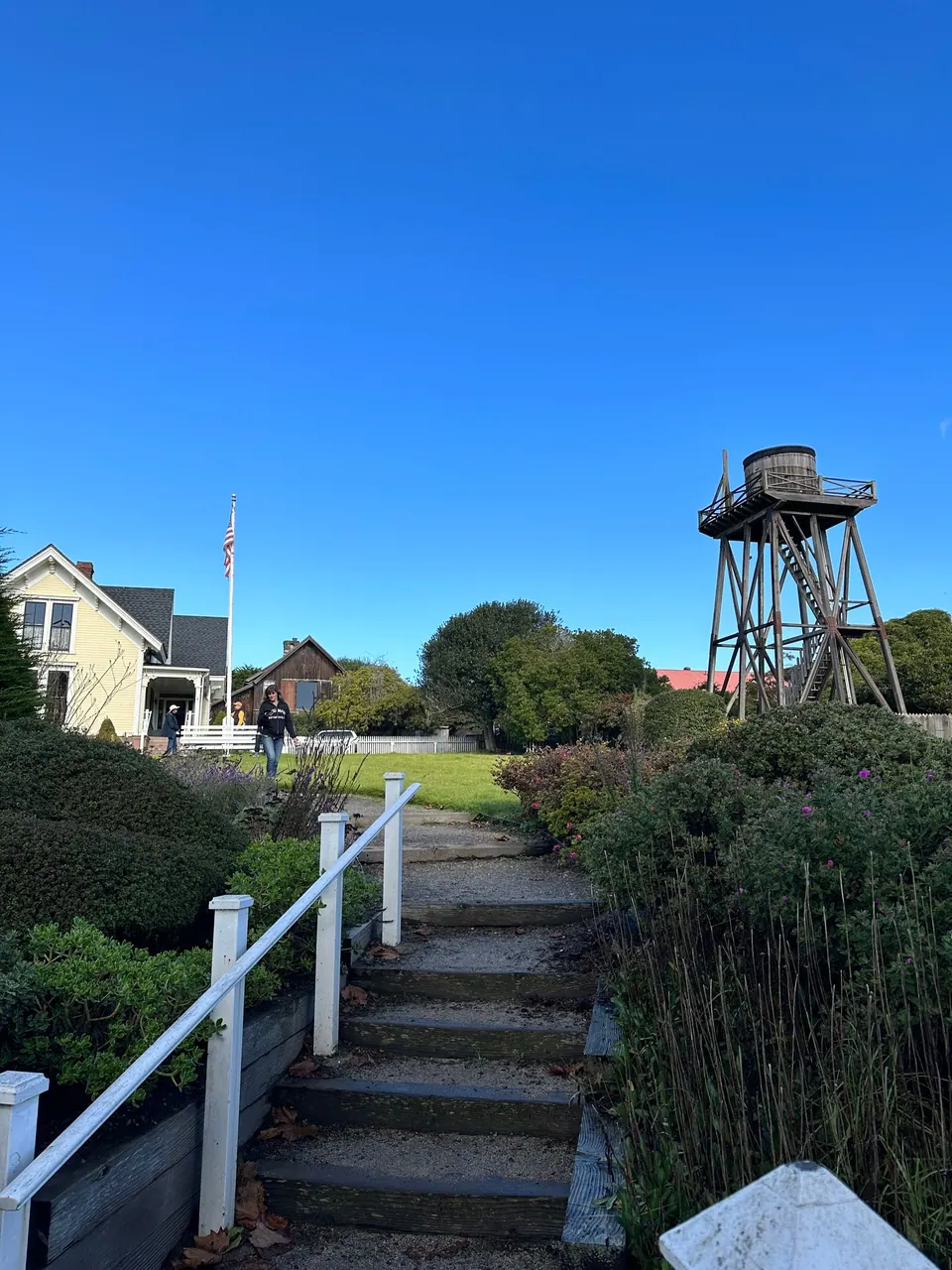 A wooden water tower rises over a white house and a garden with a stepped path and a white handrail.