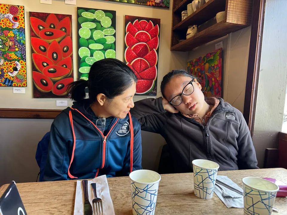 A woman in a blue jacket and a girl with glasses in a gray fleece sit at a corner table under paintings of red and green fruit, with three empty paper cups.