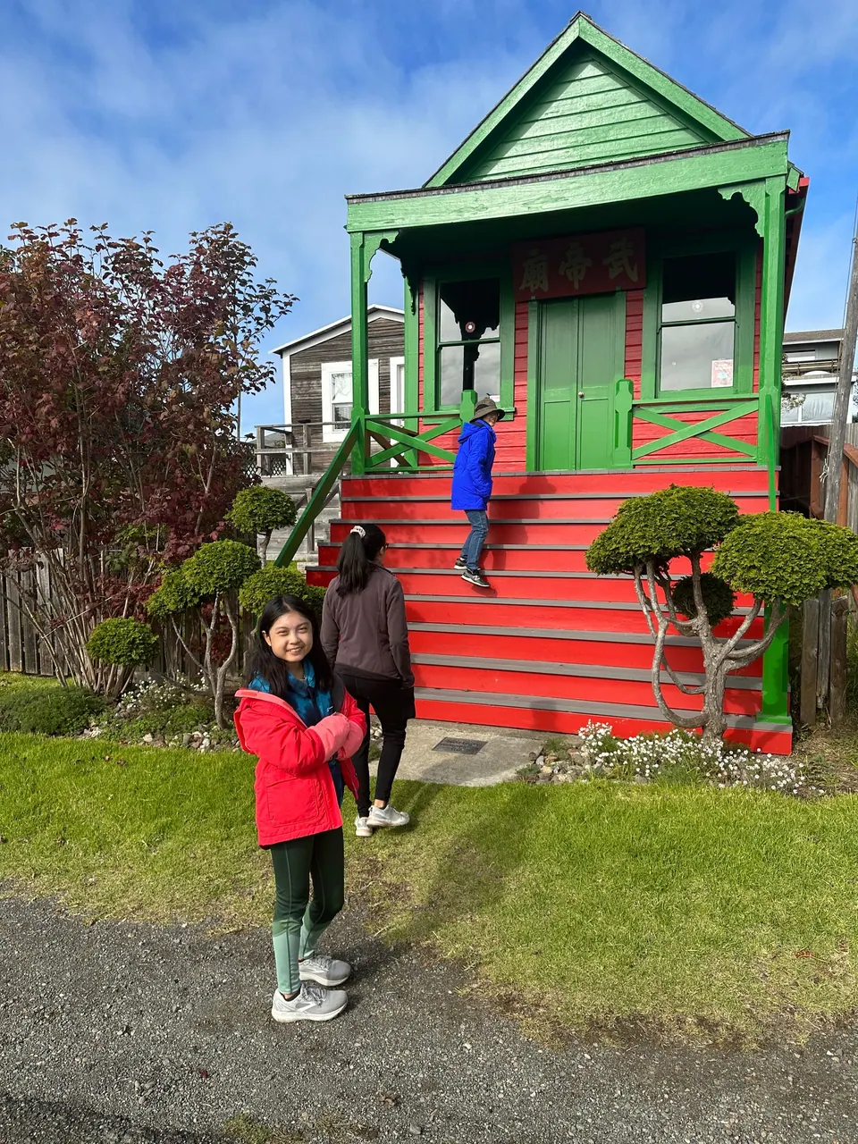 A girl in a red jacket stands on the grass in front of a small temple building with green trim and red steps, two people climbing the steps.